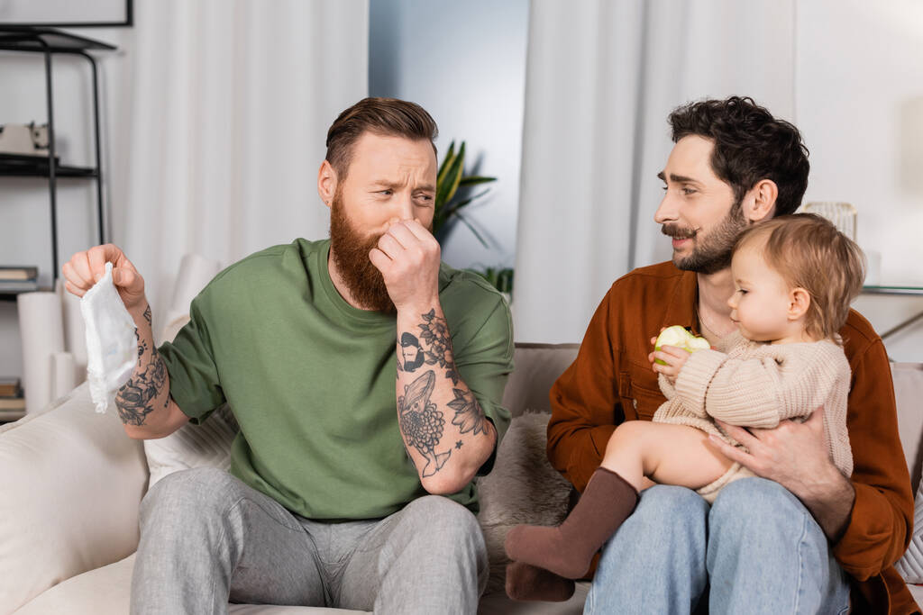 walgelijke gay man plugging neus terwijl het houden van luier in de buurt partner met baby dochter thuis  - Foto, afbeelding