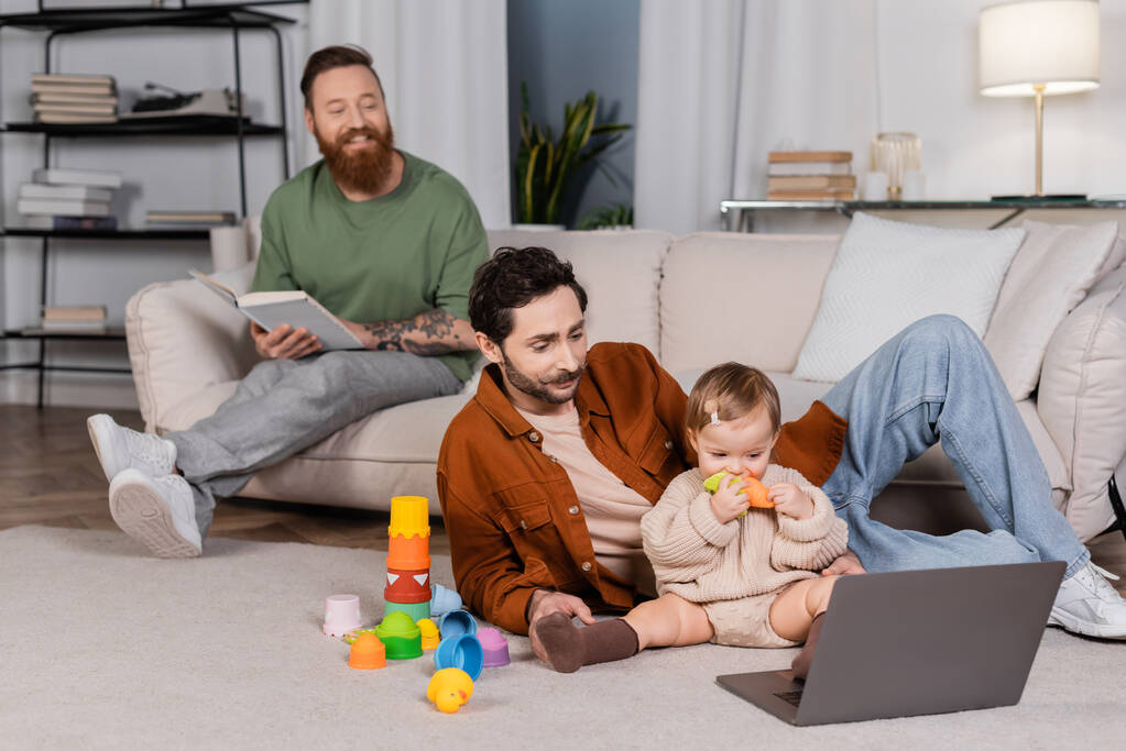 Baby daughter holding toy near laptop and fathers at home  - Photo, image