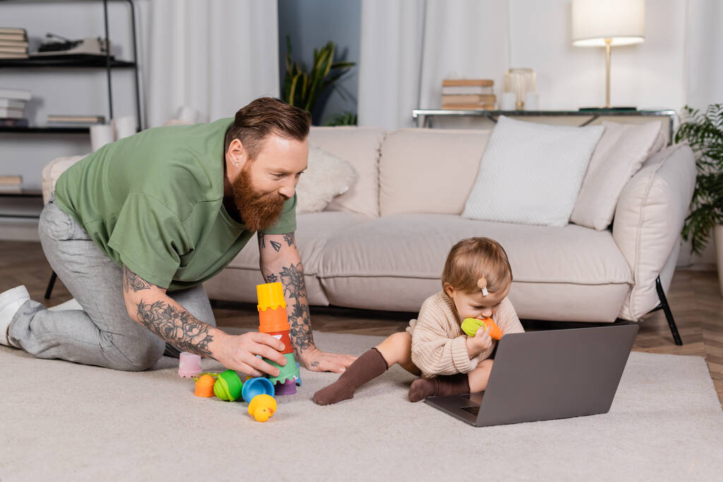 Tattooed father holding toys near baby daughter and laptop at home  - 写真・画像
