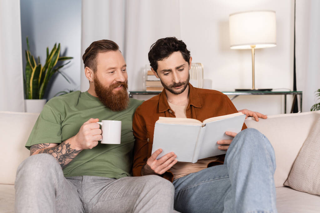 glimlachende gay man holding kopje koffie terwijl partner lezen boek thuis  - Foto, afbeelding