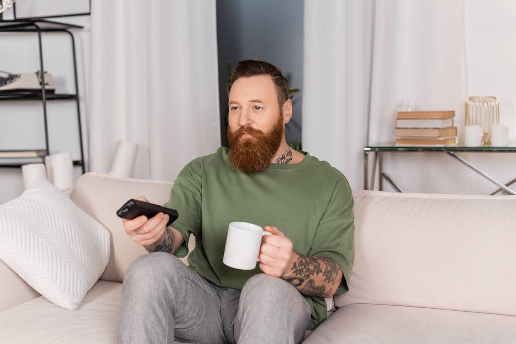 Татуйований чоловік тримає чашку кави і дивиться телевізор на дивані вдома
  - Фото, зображення