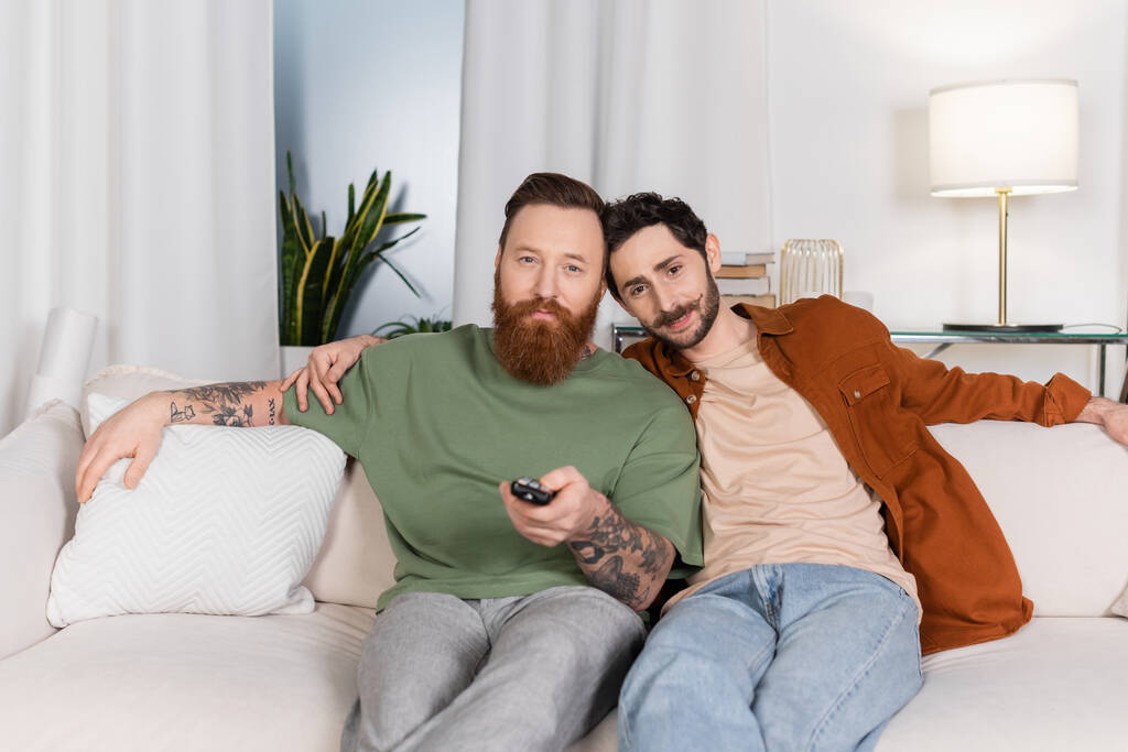 Bearded same sex couple watching tv on couch at home  - Foto, Imagem