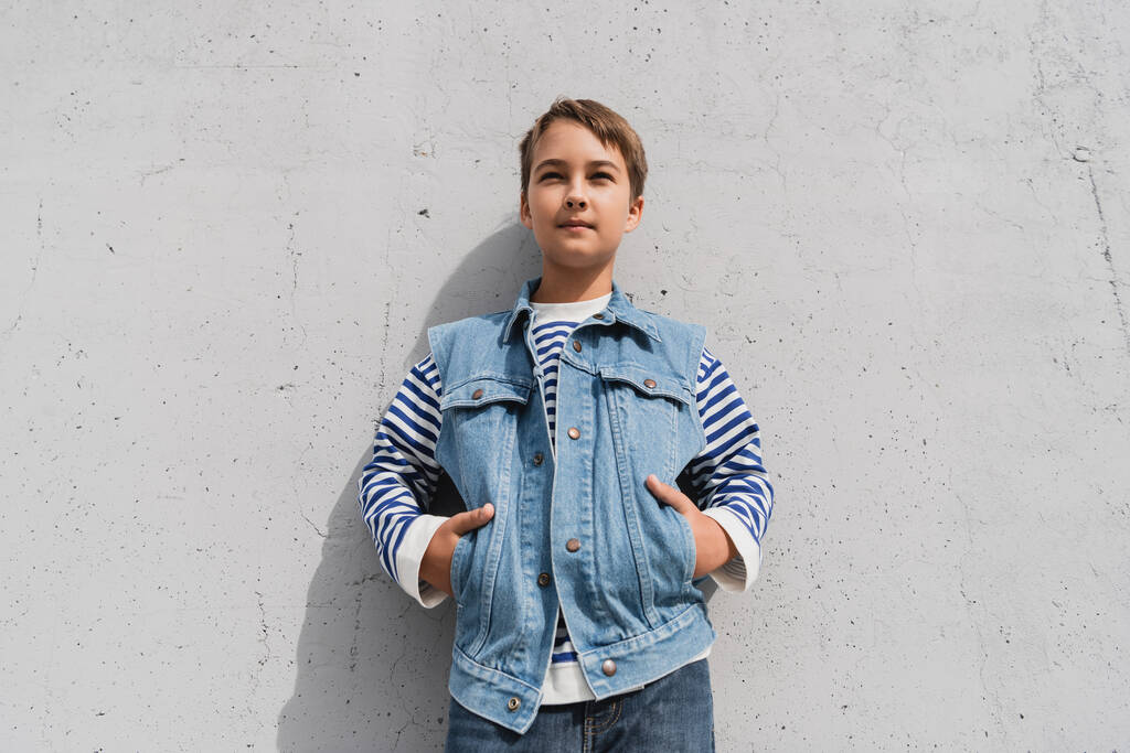 stylish preteen boy in denim vest and striped long sleeve shirt posing with hands in pockets near mall with grey wall - Valokuva, kuva