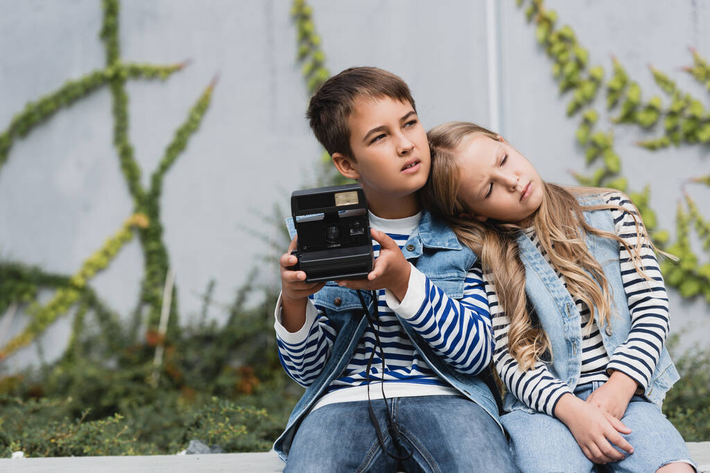 menina em colete jeans e jeans apoiados no ombro do menino pré-adolescente em roupas elegantes segurando câmera vintage  - Foto, Imagem