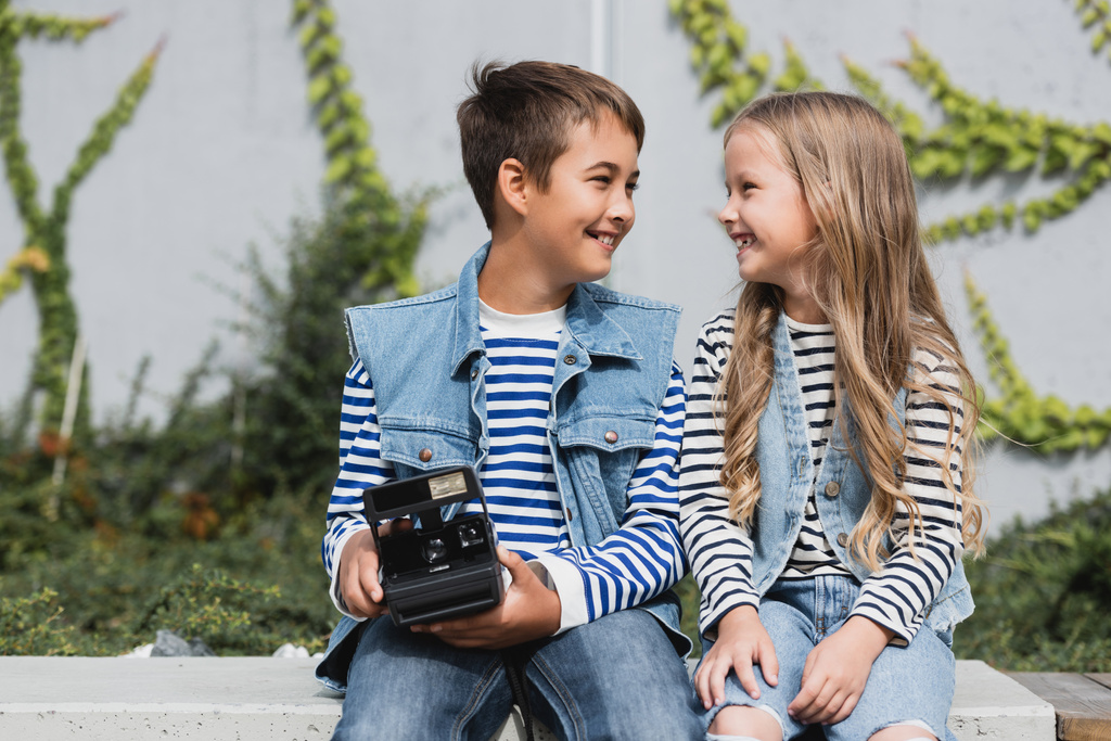 šťastný před dospívající chlapec ve stylovém oblečení držení vintage kamera v blízkosti usmívající se dívka  - Fotografie, Obrázek