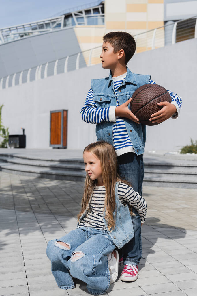 girl in stylish clothes hugging legs of boy with basketball standing near mall  - Φωτογραφία, εικόνα