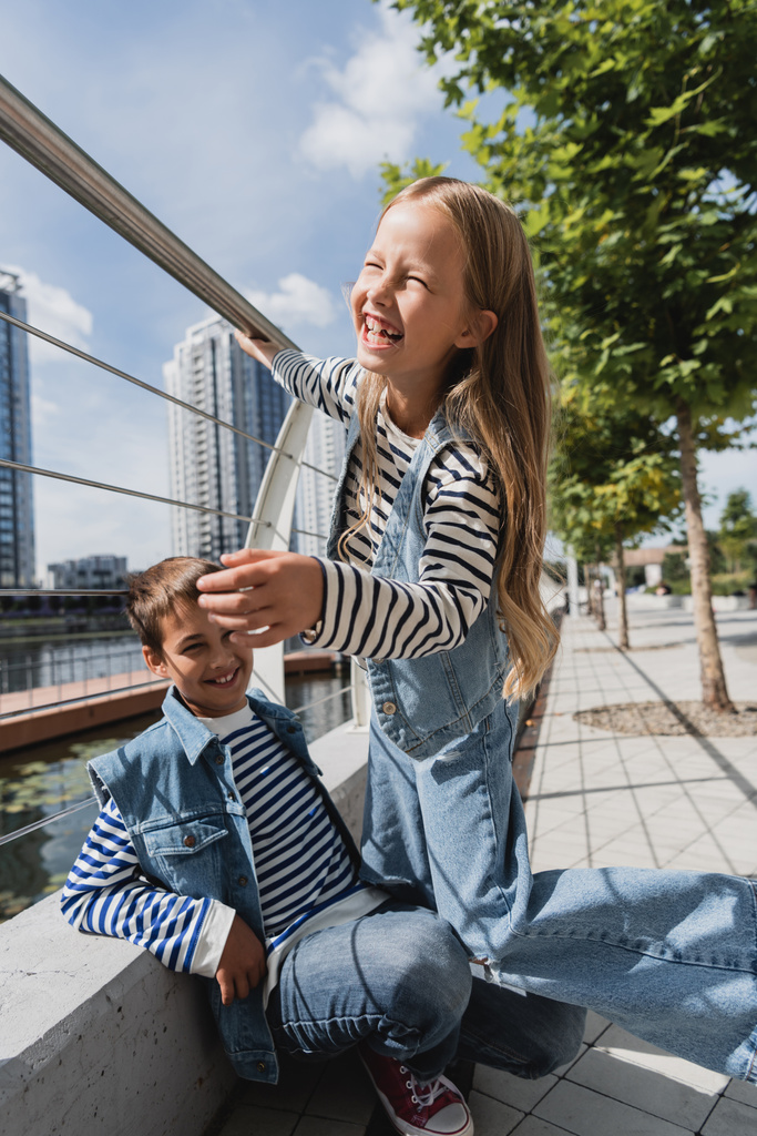 Радостная девушка в джинсовом жилете и джинсах смеется рядом с мальчиком рядом с металлическим забором на берегу реки - Фото, изображение