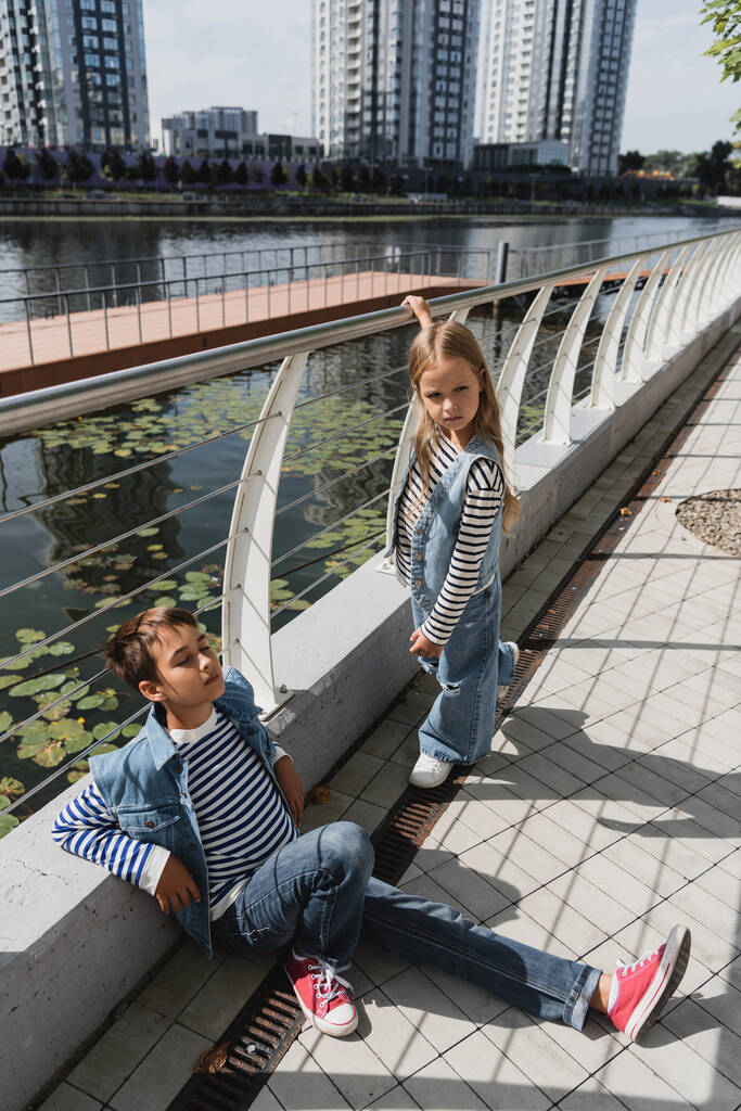 Blick aus der Vogelperspektive auf gut gekleidete Kinder in Jeanswesten und Jeans, die in der Nähe eines Metallzauns am Ufer des Flusses posieren  - Foto, Bild