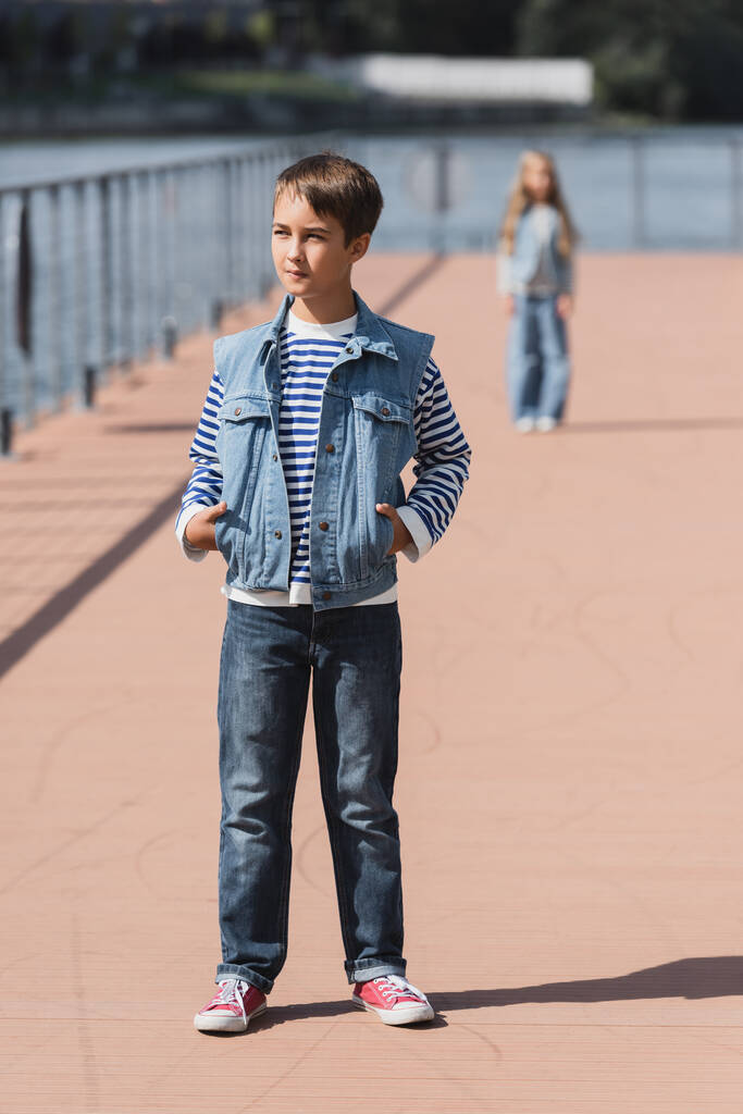 full length of preteen boy in stylish outfit posing with hands in pockets near blurred girl on river embankment  - Zdjęcie, obraz