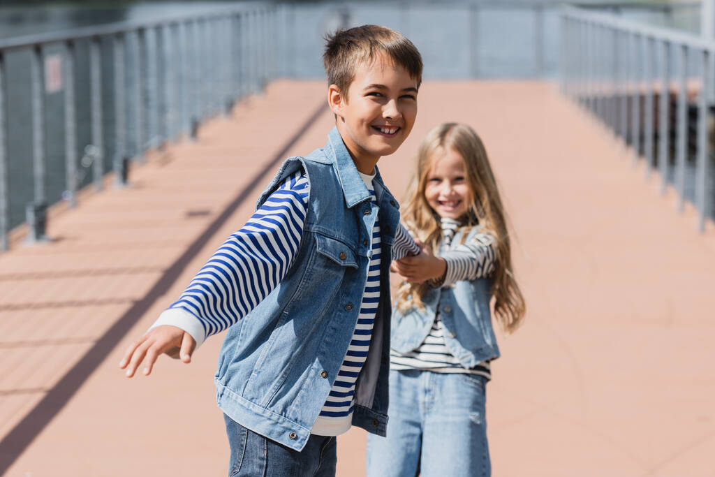 щасливі діти в джинсовому одязі тримаються за руки, розважаючись на набережній річки
  - Фото, зображення