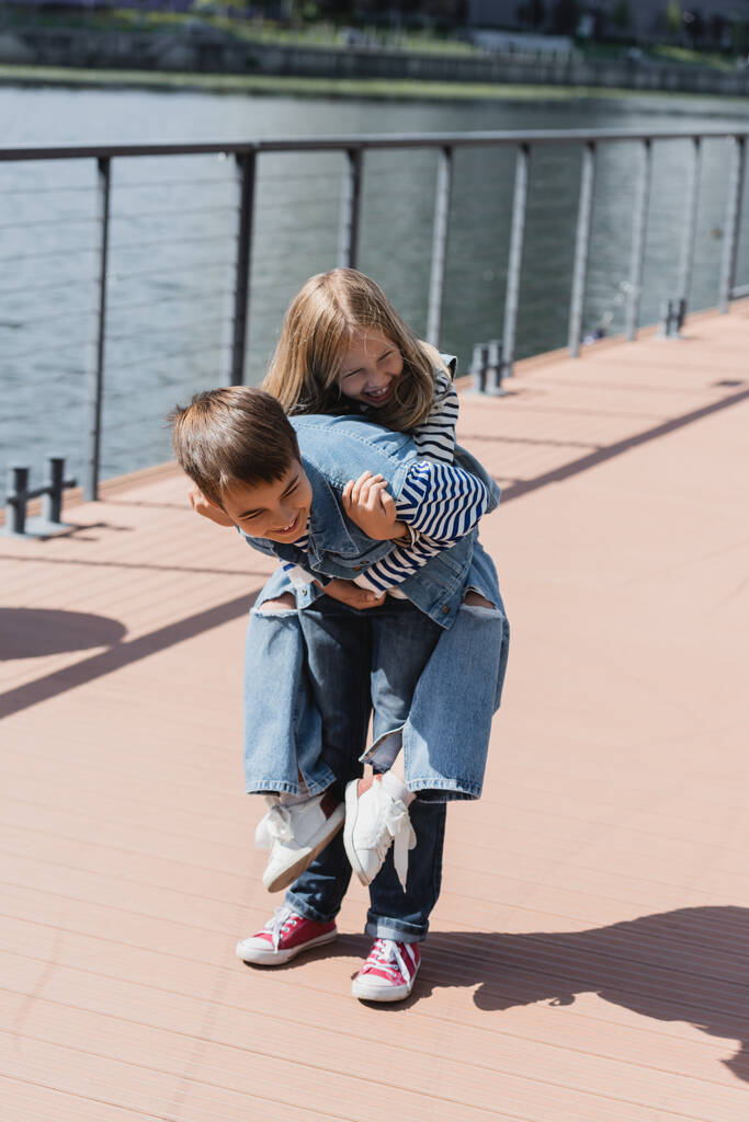 happy preteen boy in denim outfit piggybacking smiling girl on river embankment  - Фото, изображение