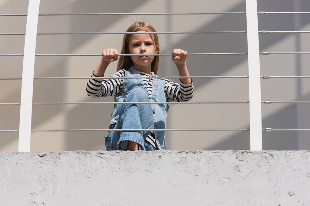 金属製のフェンスの近くのカメラを見てデニムの服でスタイリッシュな子供の低角度ビュー  - 写真・画像