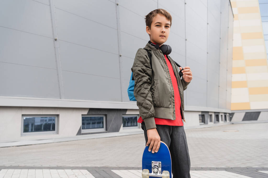 preteen αγόρι στο σακάκι βομβαρδιστικό κρατώντας penny σκάφους, ενώ στέκεται κοντά στο εμπορικό κέντρο  - Φωτογραφία, εικόνα