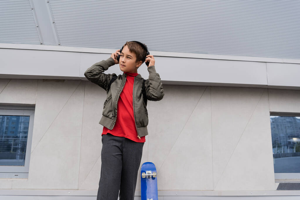 preteen boy in bomber jacket wearing wireless headphones near penny board while standing near mall  - Фото, изображение