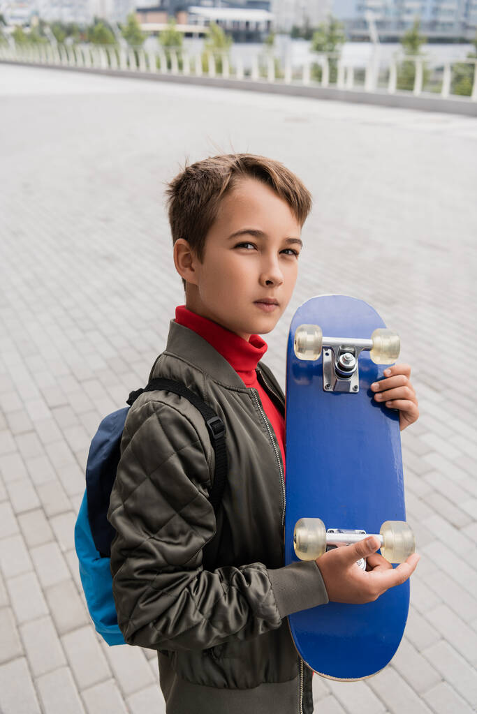preteen boy in trendy bomber jacket standing with backpack while holding penny board  - Fotó, kép
