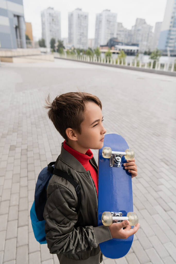 vista lateral del niño preadolescente en la chaqueta de bomber de moda de pie con la mochila mientras sostiene penny board  - Foto, Imagen