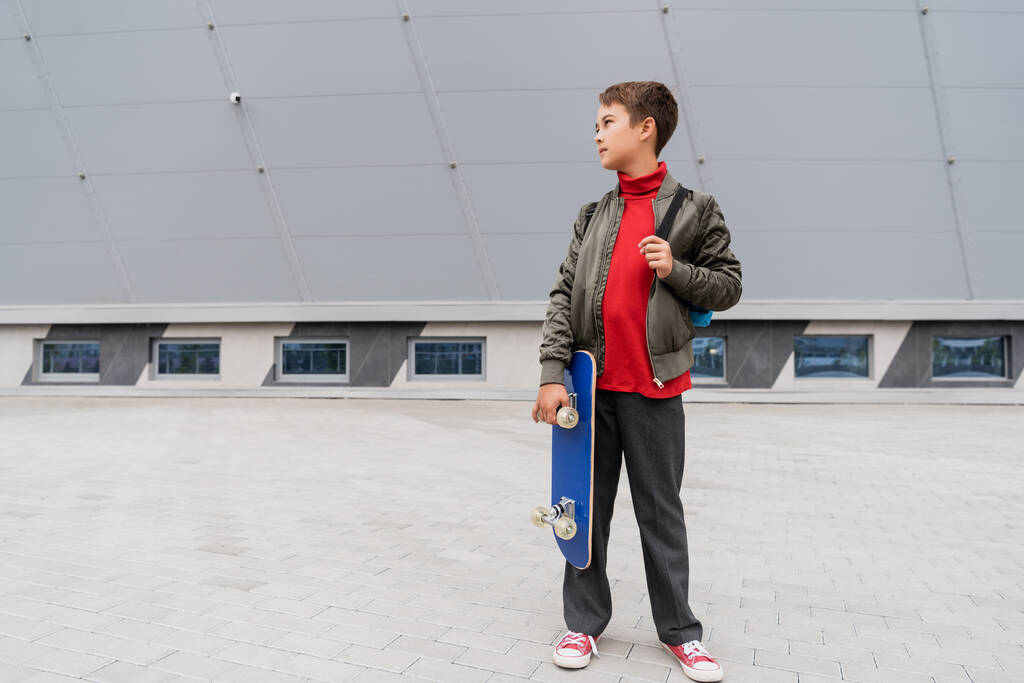 full length of preteen boy in trendy bomber jacket holding penny board near mall building  - Foto, Imagen