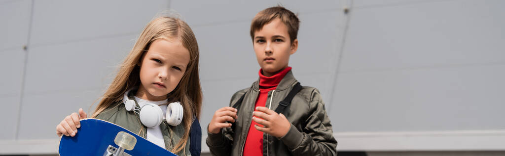 niña preadolescente en auriculares inalámbricos que sostienen penny board mientras está de pie con el niño cerca del centro comercial, pancarta  - Foto, Imagen