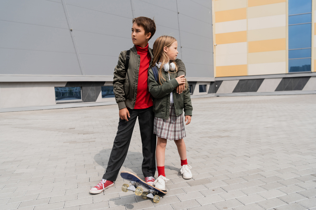 full length of preteen boy in stylish bomber jacket hugging girl in skirt while standing near penny board  - Photo, Image