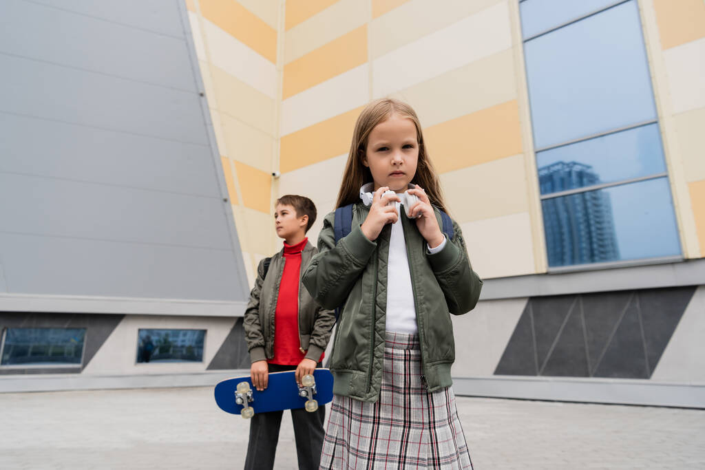 preteen girl in wireless headphones standing with stylish boy holding penny board on blurred background  - Zdjęcie, obraz