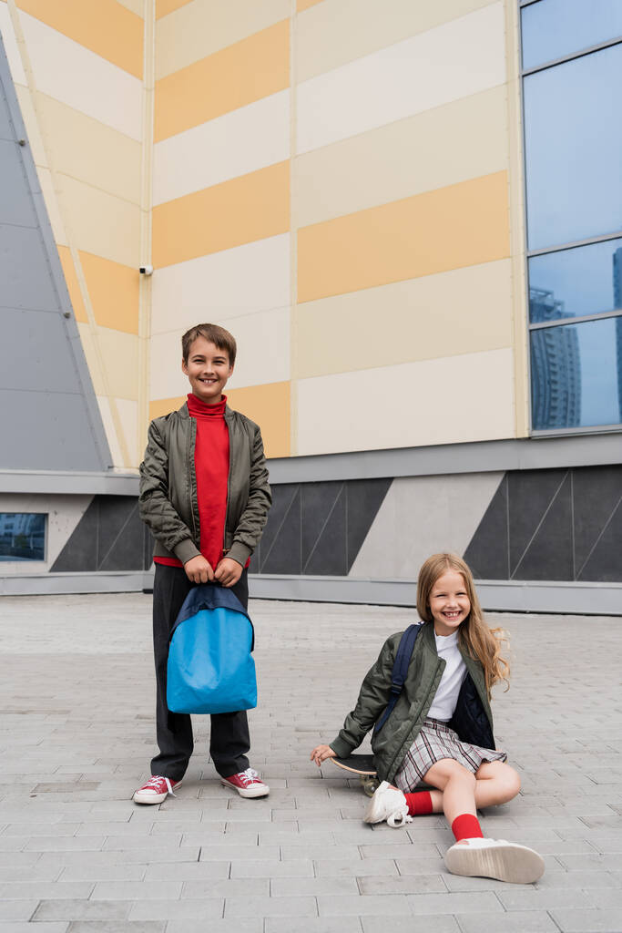 chica feliz en falda sentado en penny board al lado de chico con estilo con mochila cerca del centro comercial  - Foto, imagen
