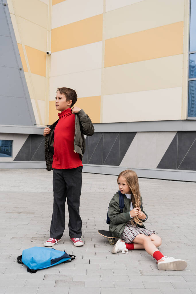 preteen girl in skirt sitting on penny board next to stylish boy wearing bomber jacket near mall  - 写真・画像