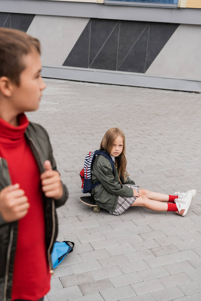 preteen girl in skirt sitting on penny board near stylish boy on blurred foreground  - 写真・画像