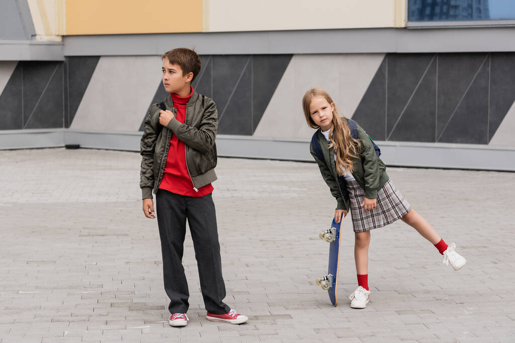 pleine longueur de fille préadolescente en jupe tenant penny conseil près du garçon en veste bombardier tout en se tenant à côté du centre commercial  - Photo, image