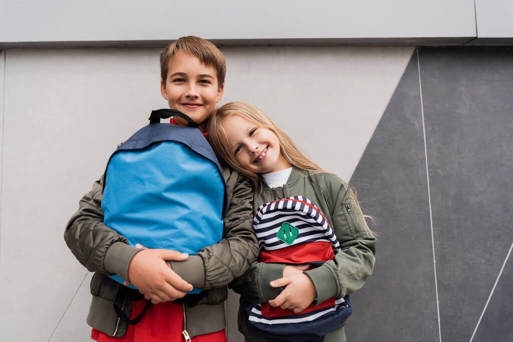 low angle view of happy kids in trendy bomber jackets holding backpacks while standing near mall  - Fotoğraf, Görsel
