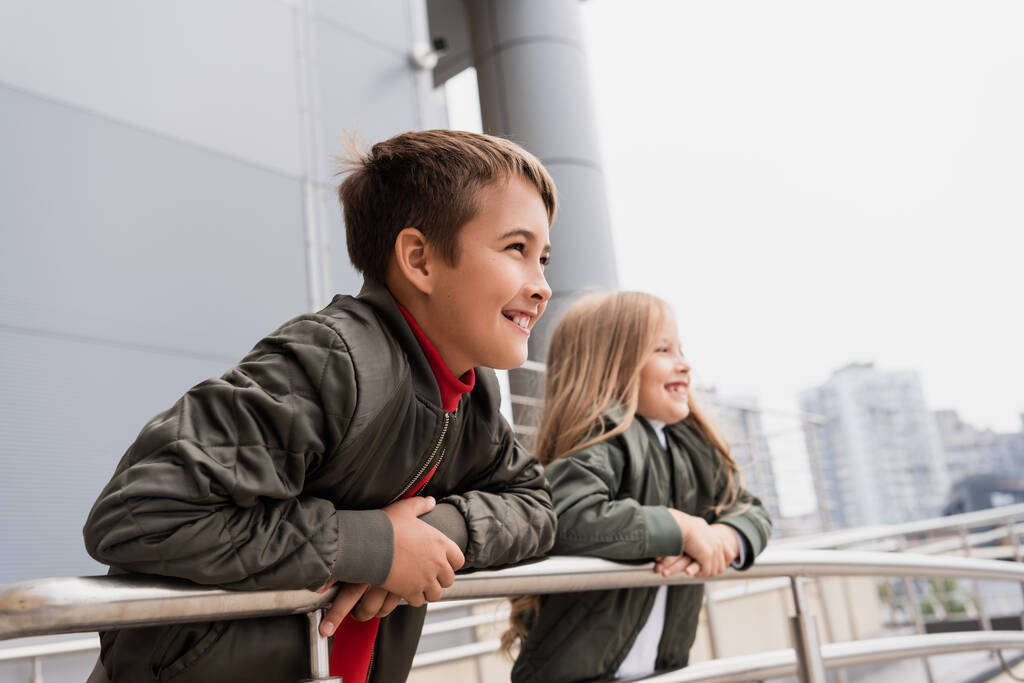 happy preteen kids in bomber jackets leaning on metallic handrails near mall  - Zdjęcie, obraz