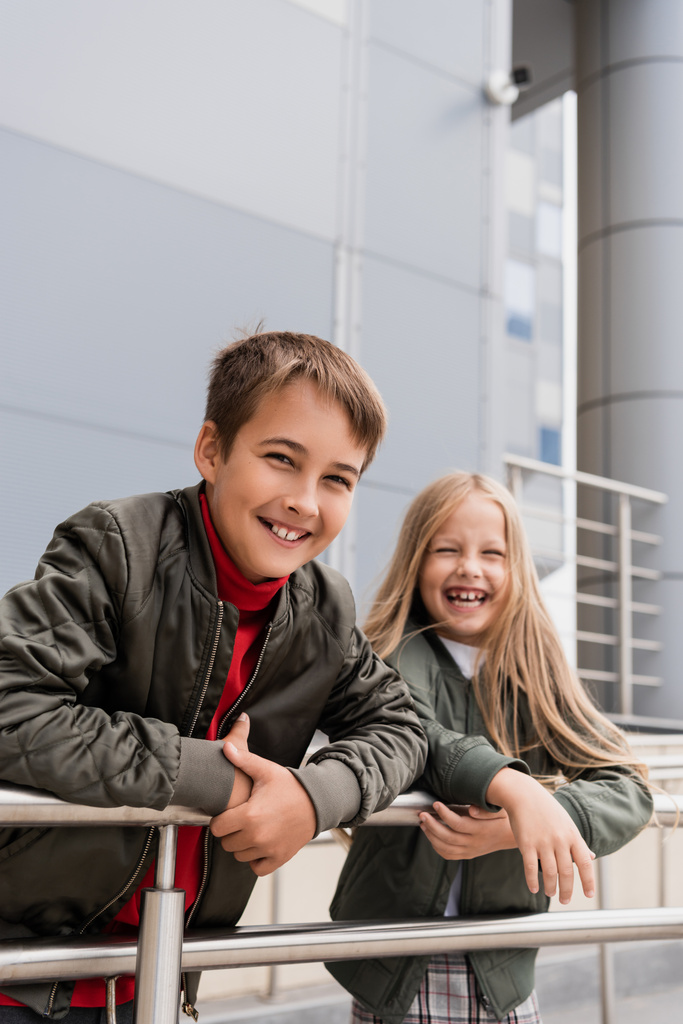 cheerful preteen kids in bomber jackets leaning on metallic handrails near mall  - 写真・画像