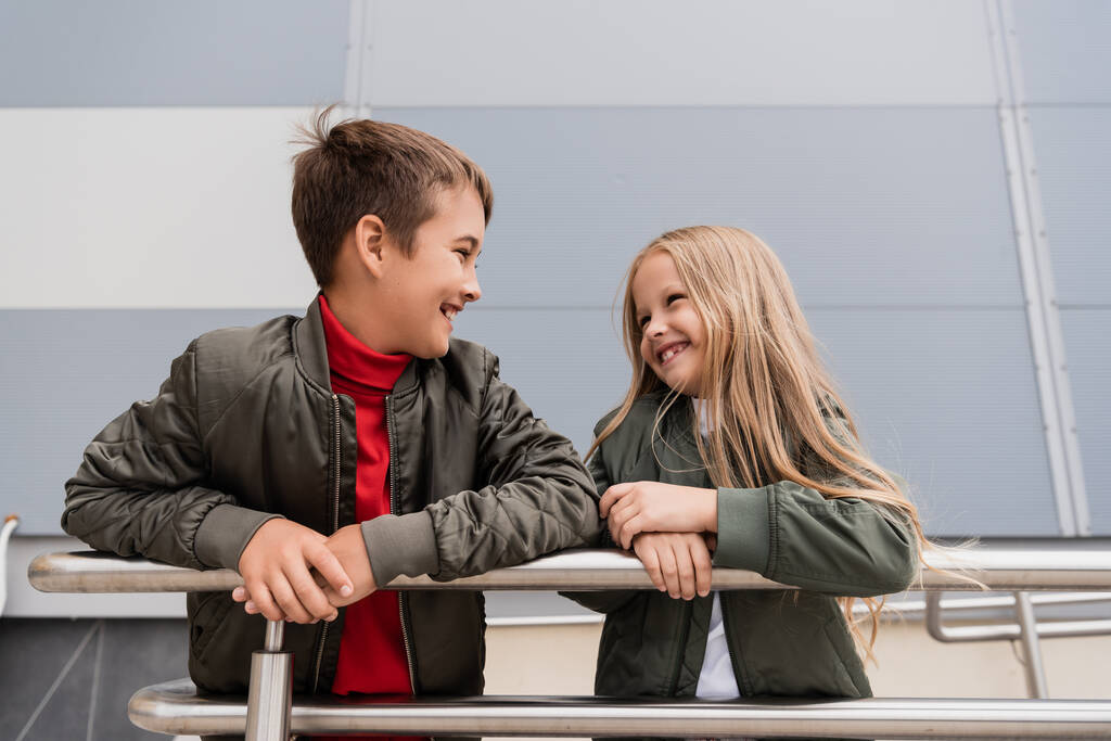 happy preteen kids in bomber jackets looking at each other while leaning on metallic handrails near mall  - Fotografie, Obrázek