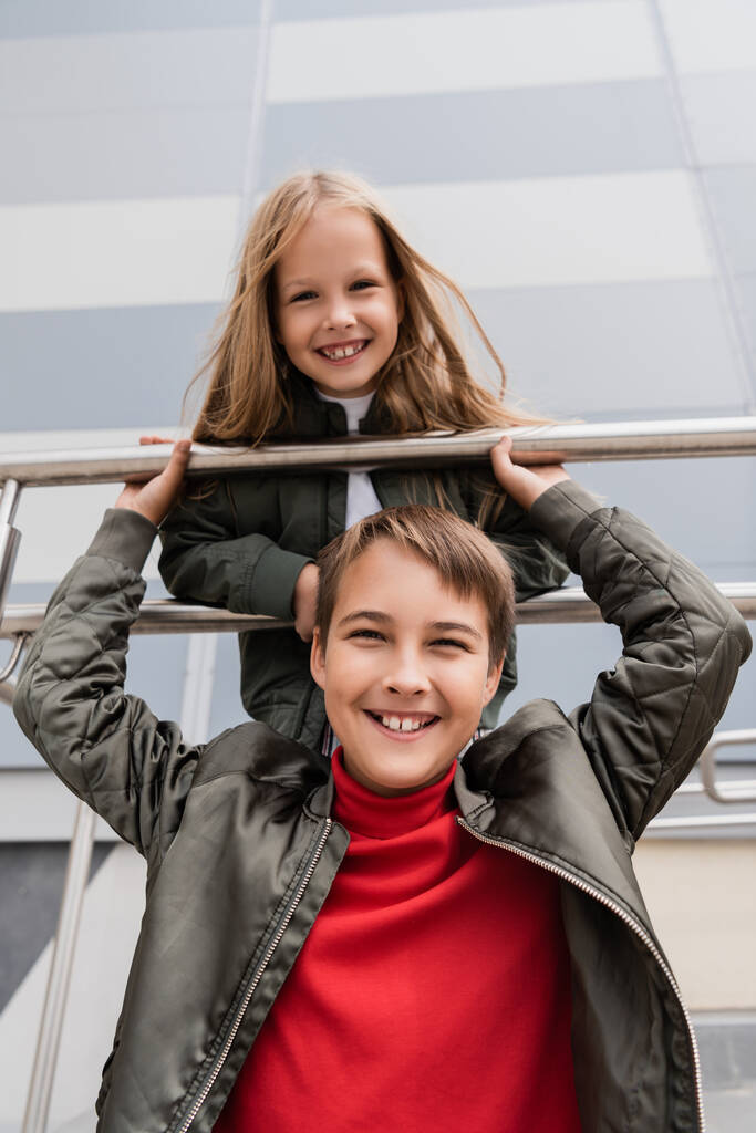 low angle view of happy and well dressed preteen kids in bomber jackets leaning on metallic handrails near mall  - Photo, Image