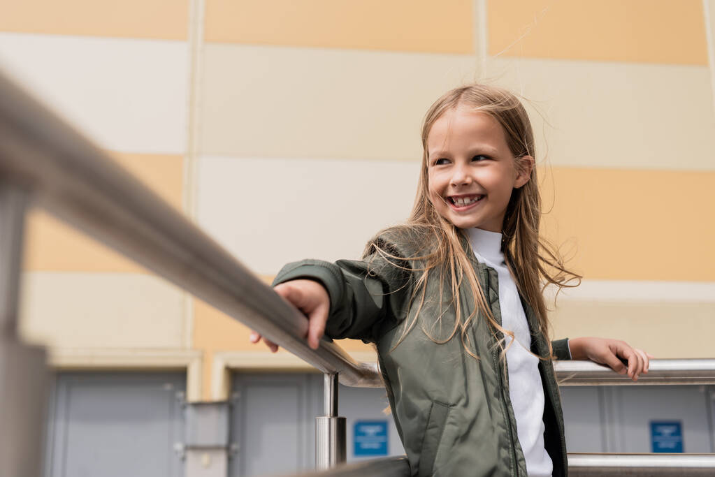 spokojený dospívající dívka ve stylové bombardér bunda opírající se o kovové madla v blízkosti nákupního centra  - Fotografie, Obrázek
