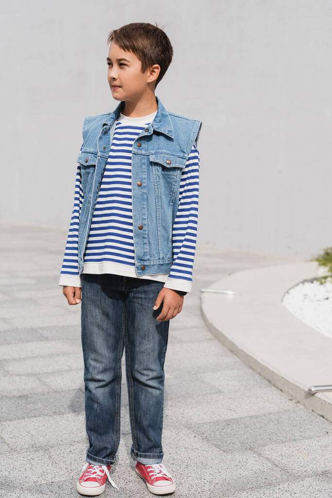 longitud completa de niño bien vestido en camisa de manga larga a rayas y chaleco de mezclilla de pie al aire libre  - Foto, Imagen