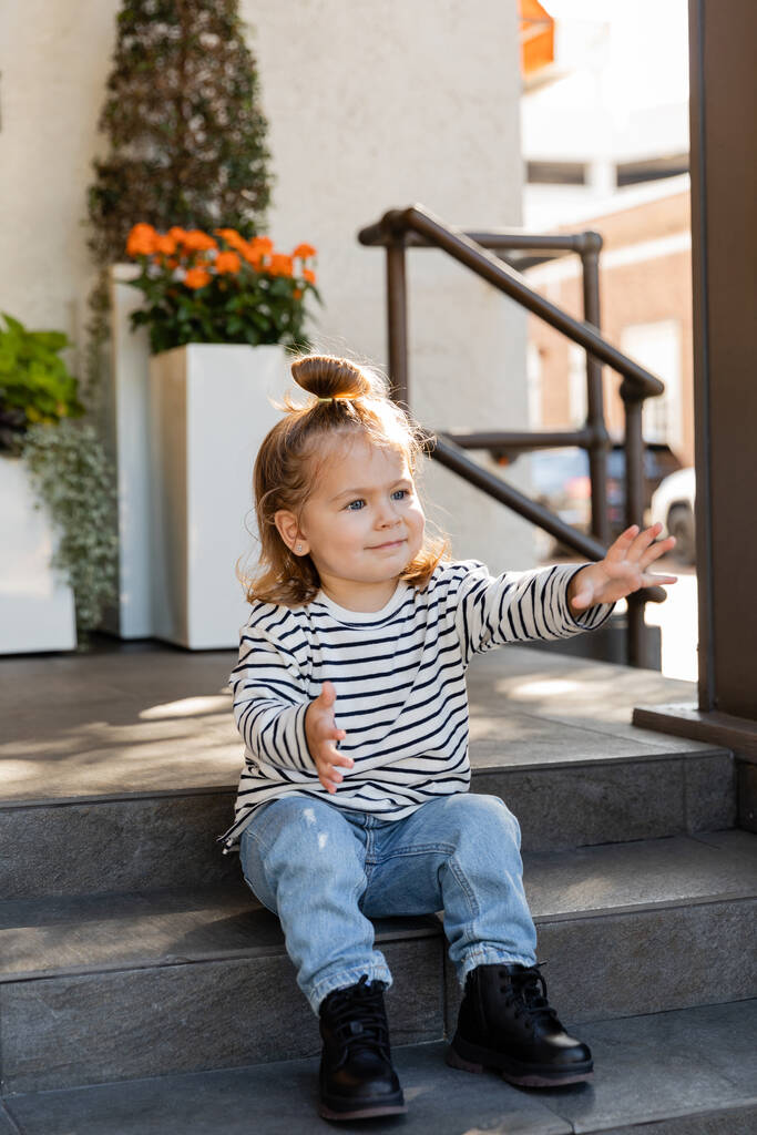Kleinkind in lässiger Kleidung fordert Aufmerksamkeit und gestikuliert, während es auf einer Treppe in der Nähe des Hauses sitzt  - Foto, Bild