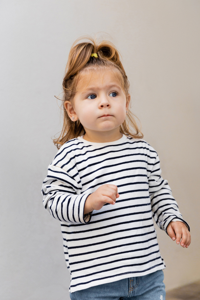 Малышка в полосатой рубашке с длинным рукавом и джинсах смотрит в сторону  - Фото, изображение