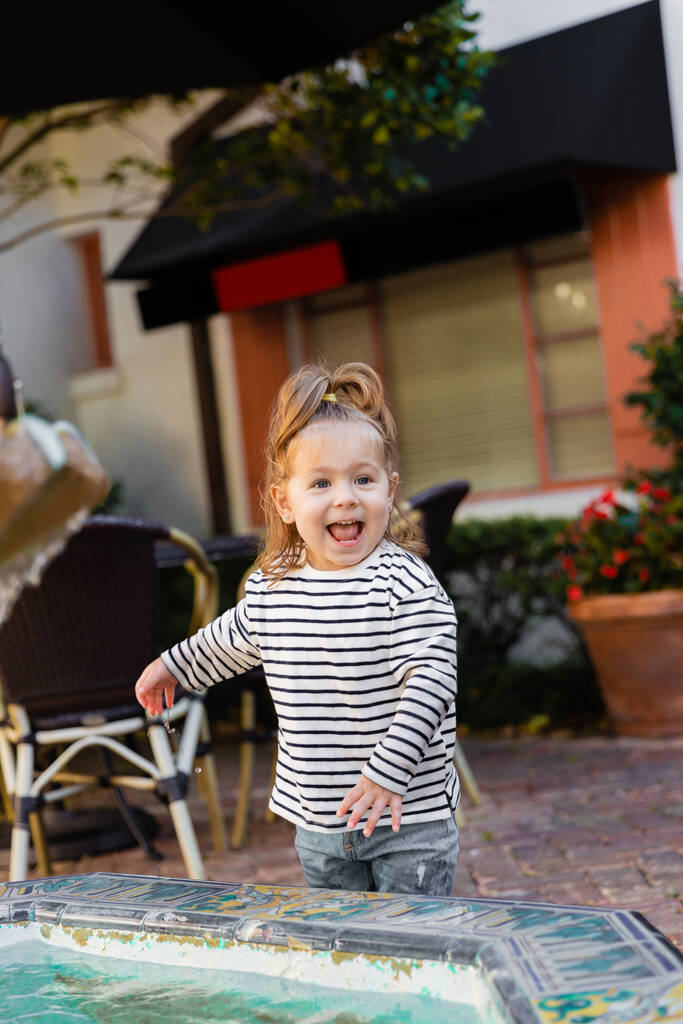 gioiosa bambina in camicia a maniche lunghe a righe in piedi vicino alla fontana di Miami  - Foto, immagini