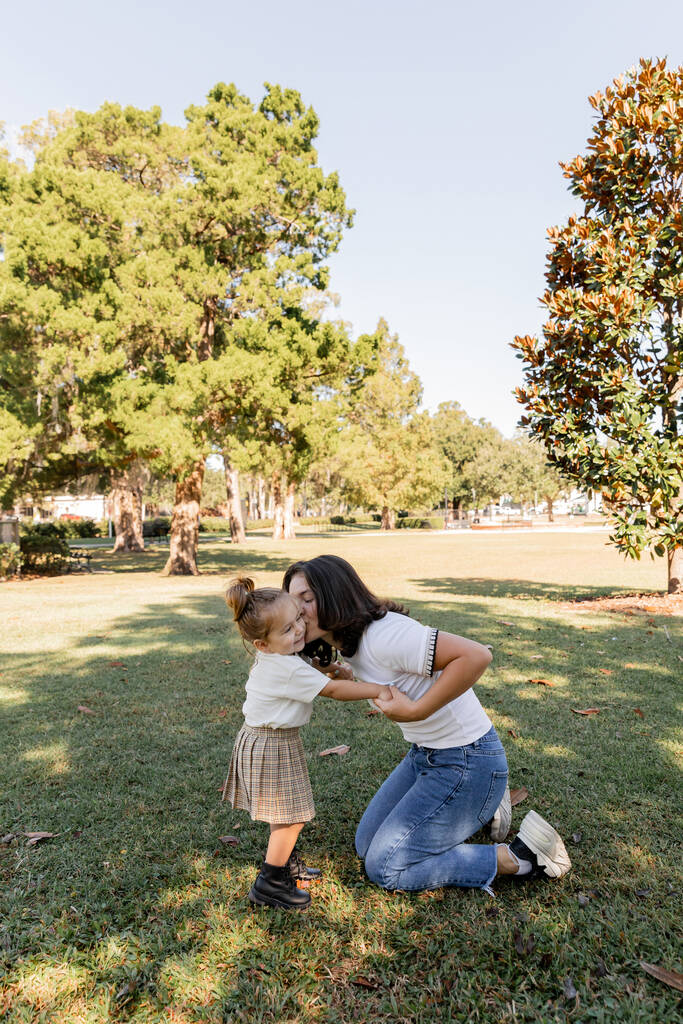 bruna madre in piedi sulle ginocchia e baciare la guancia della figlia del bambino nel parco di Miami  - Foto, immagini