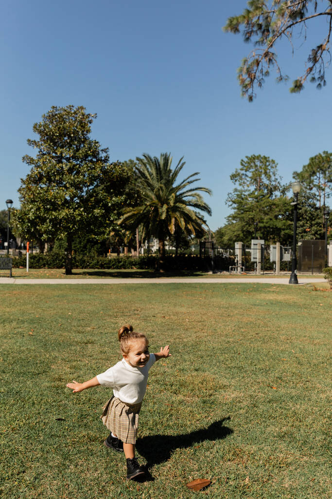 positive toddler girl standing with outstretched hands while playing in green park  - Photo, Image