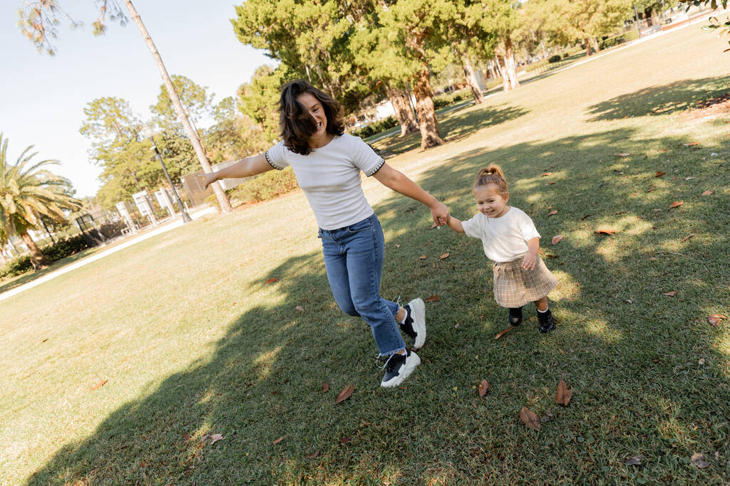緑のマイアミパークで一緒に遊んでいる間、楽しい母親と子供の手を握って  - 写真・画像