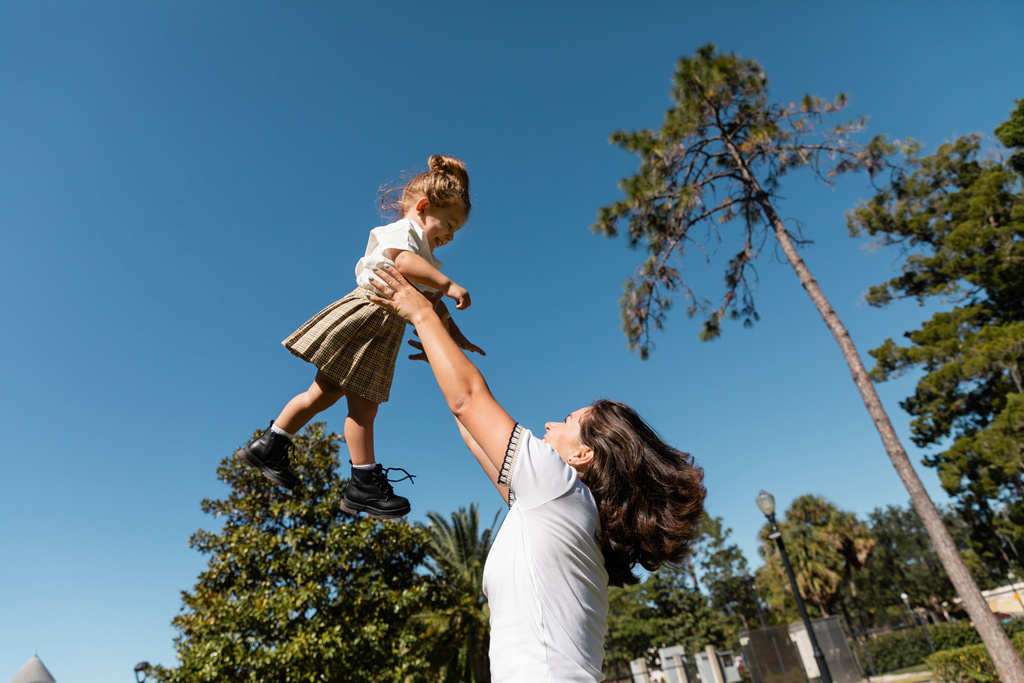 ευτυχισμένη μητέρα ανύψωση κόρη νηπίων ενάντια στα πράσινα δέντρα και τον καθαρό ουρανό  - Φωτογραφία, εικόνα