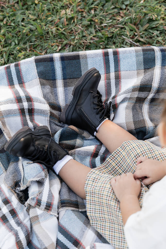 Blick von oben auf das kleine Mädchen in kariertem Rock und Stiefeln, das beim Picknick auf einer Decke sitzt  - Foto, Bild