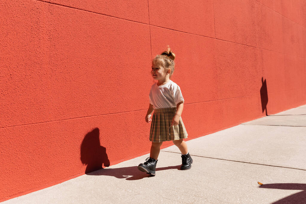 joyeuse jeune fille en jupe à carreaux et t-shirt blanc marchant près du bâtiment avec mur rouge  - Photo, image
