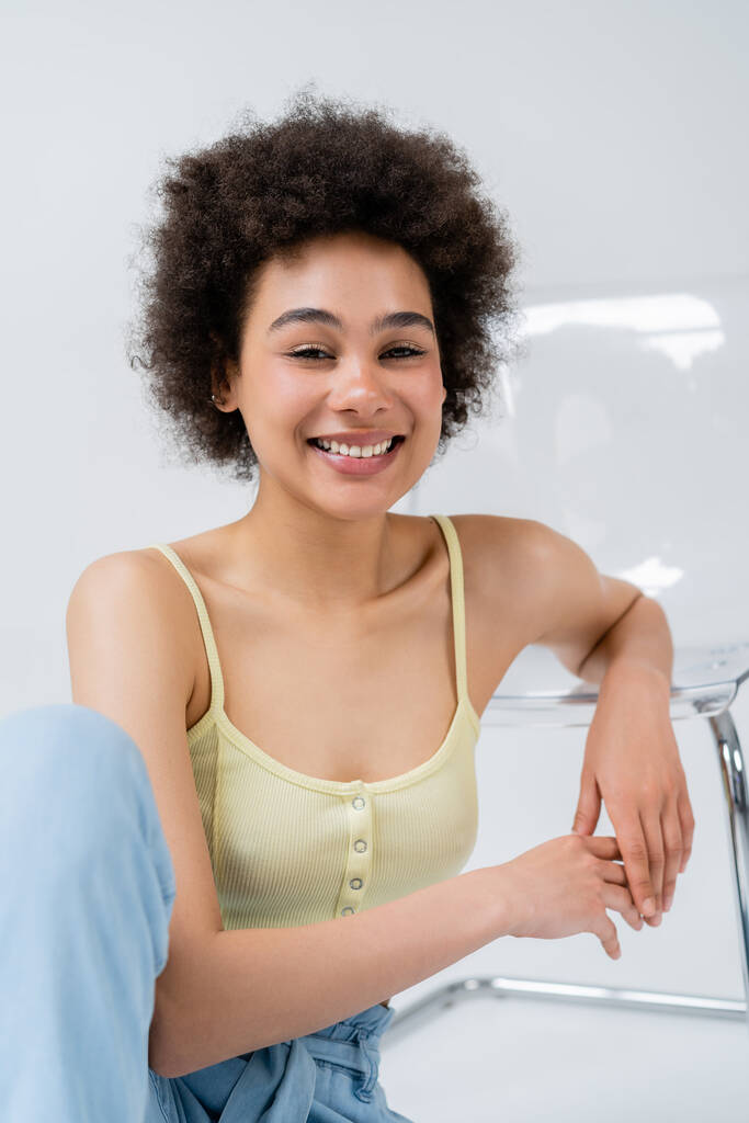 Fröhliches afrikanisch-amerikanisches Model blickt in die Kamera neben modernem Stuhl auf grauem Hintergrund  - Foto, Bild