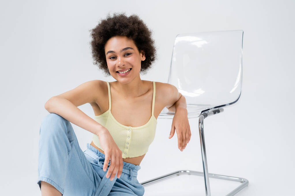 Кудрявая африканская американка сидит рядом с современным стулом на сером фоне  - Фото, изображение