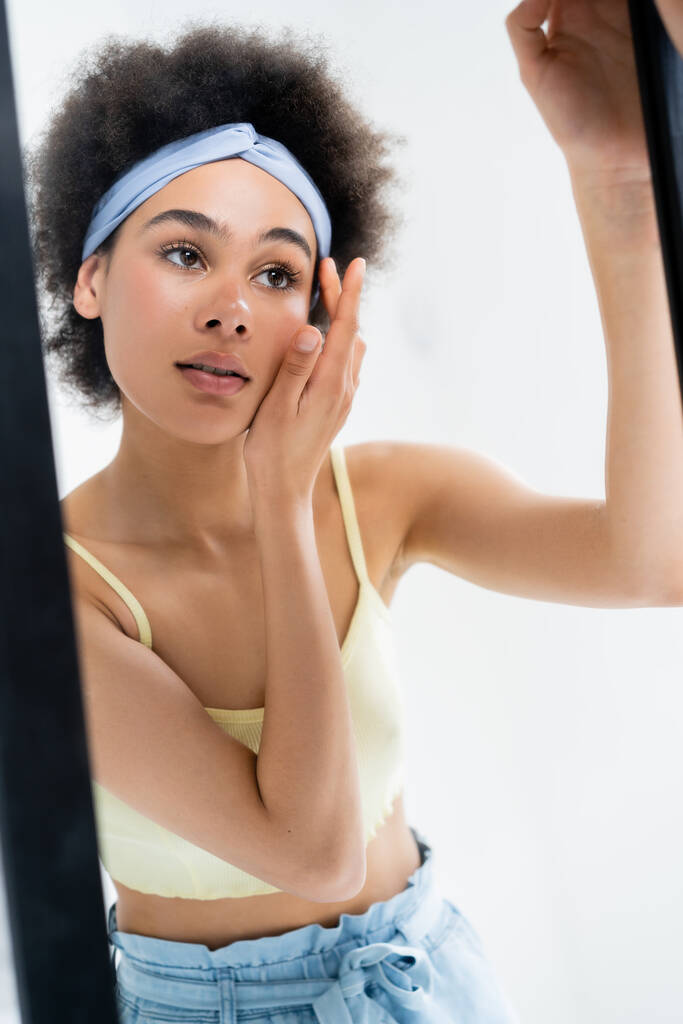 Junge Afroamerikanerin mit Stirnband berührt Gesicht in der Nähe Spiegel isoliert auf grau  - Foto, Bild