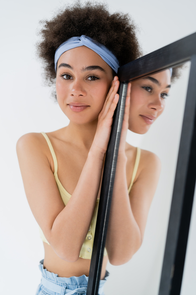 Lächelnde Afroamerikanerin mit Stirnband blickt in die Kamera in der Nähe von Spiegel isoliert auf grau  - Foto, Bild