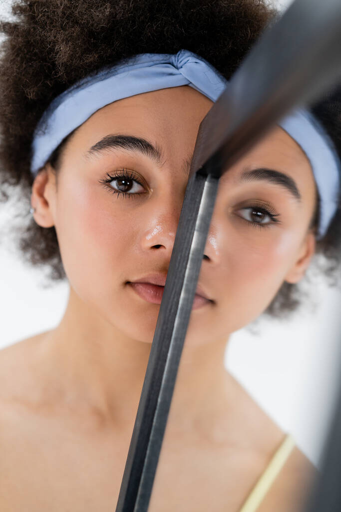 Африканская американка с повязкой на голове, смотрящая в камеру возле зеркала, изолированного на сером  - Фото, изображение