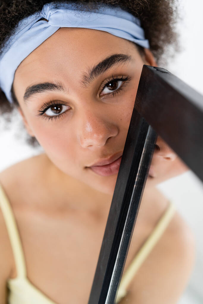 Ładna Afroamerykanka z opaską na głowie patrząca w kamerę w pobliżu zamazanego lustra odizolowanego na szaro  - Zdjęcie, obraz